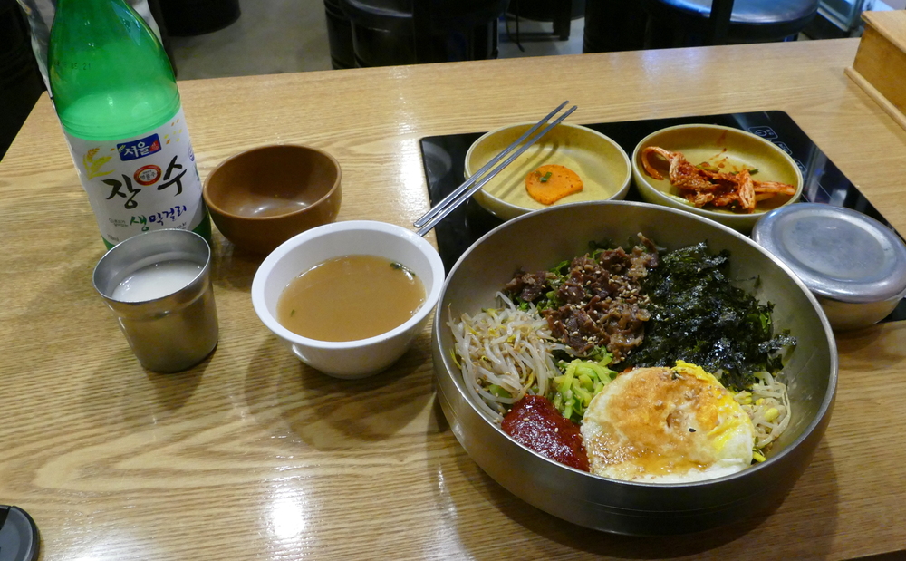 a bowl of bibimbap with a cup and bottle of makgeolli next to it