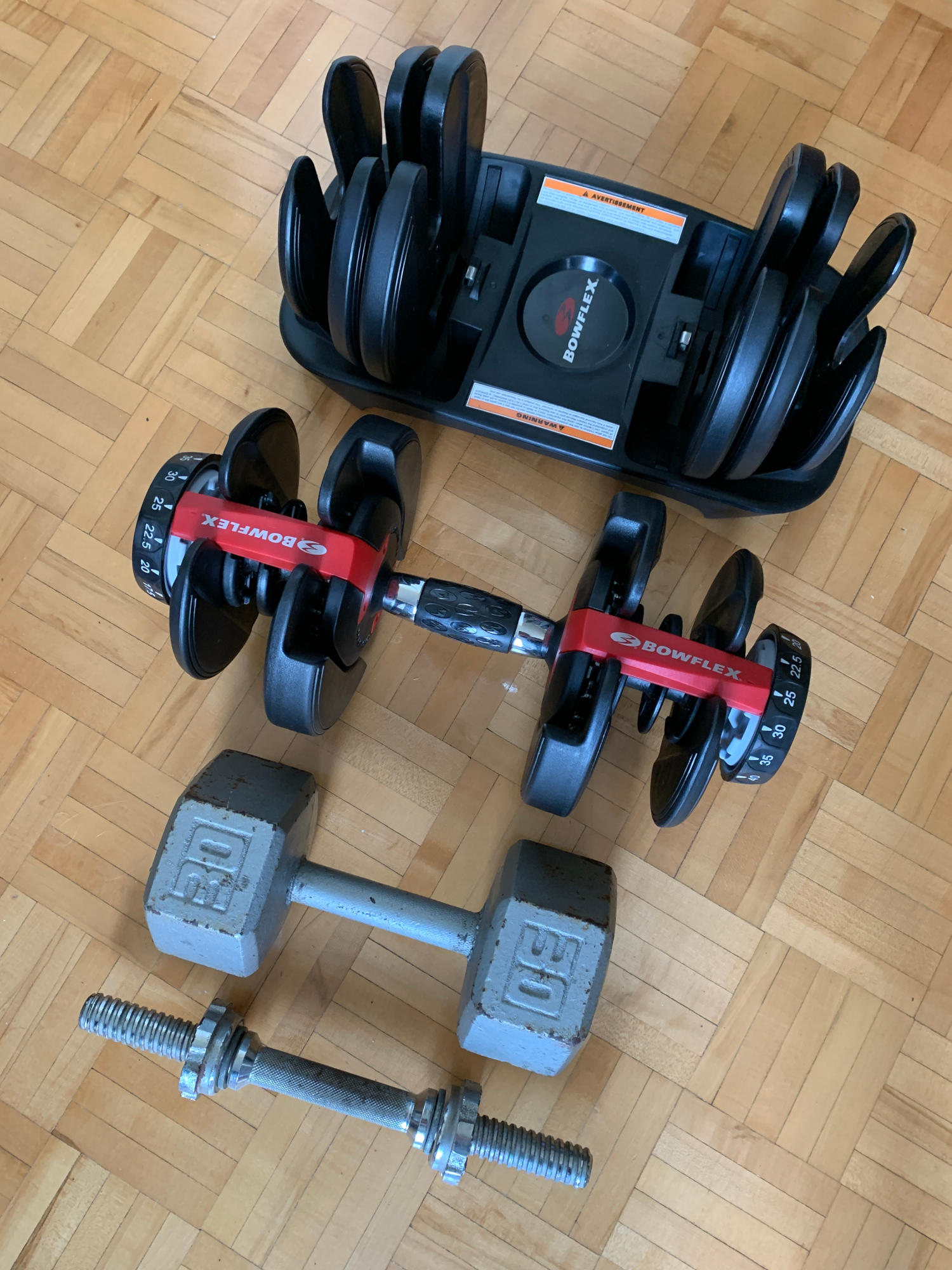 adjustable dumbbells set to 25 lb., sitting next to the tray with the remaining plates, with a 30 lb. standard dumbbell and a twist-on dumbbell spindle (both smaller) for size comparison.