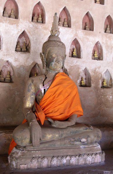 A large seated Buddha broken and held together with plastic ribbon against a wall with many small Buddhas.