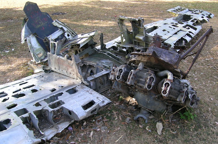 The battered remnants of the wings, seat, and engine of a fighter plane lying in a park.